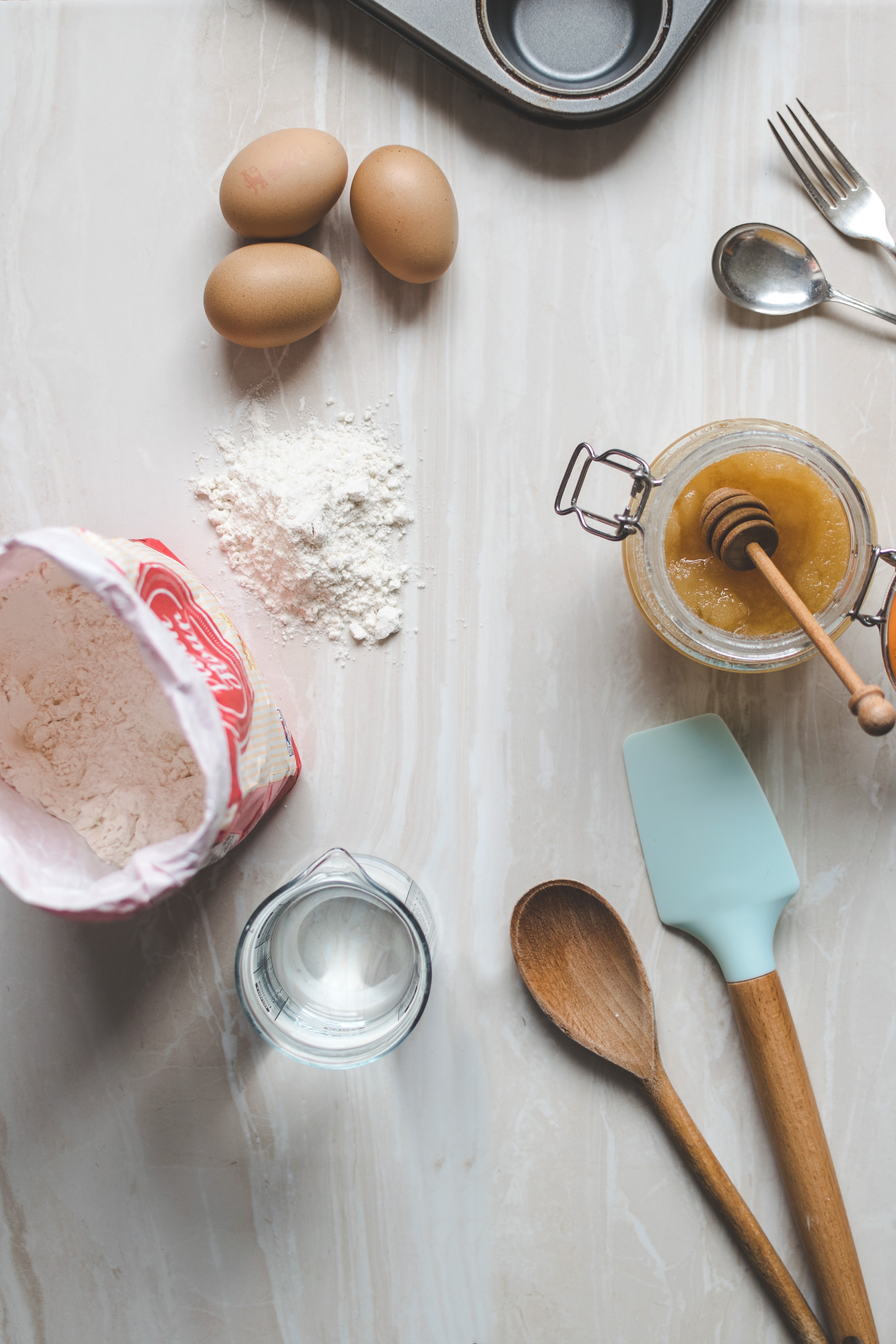 Photo of a rough mise en place, with three eggs, a honey pot, an open pack of flour, a jar with water and some spatulas. A bit of some fork, spoon and muffin tray are visible in the top corner. Keep your ingredients and tools in mind to set to work!