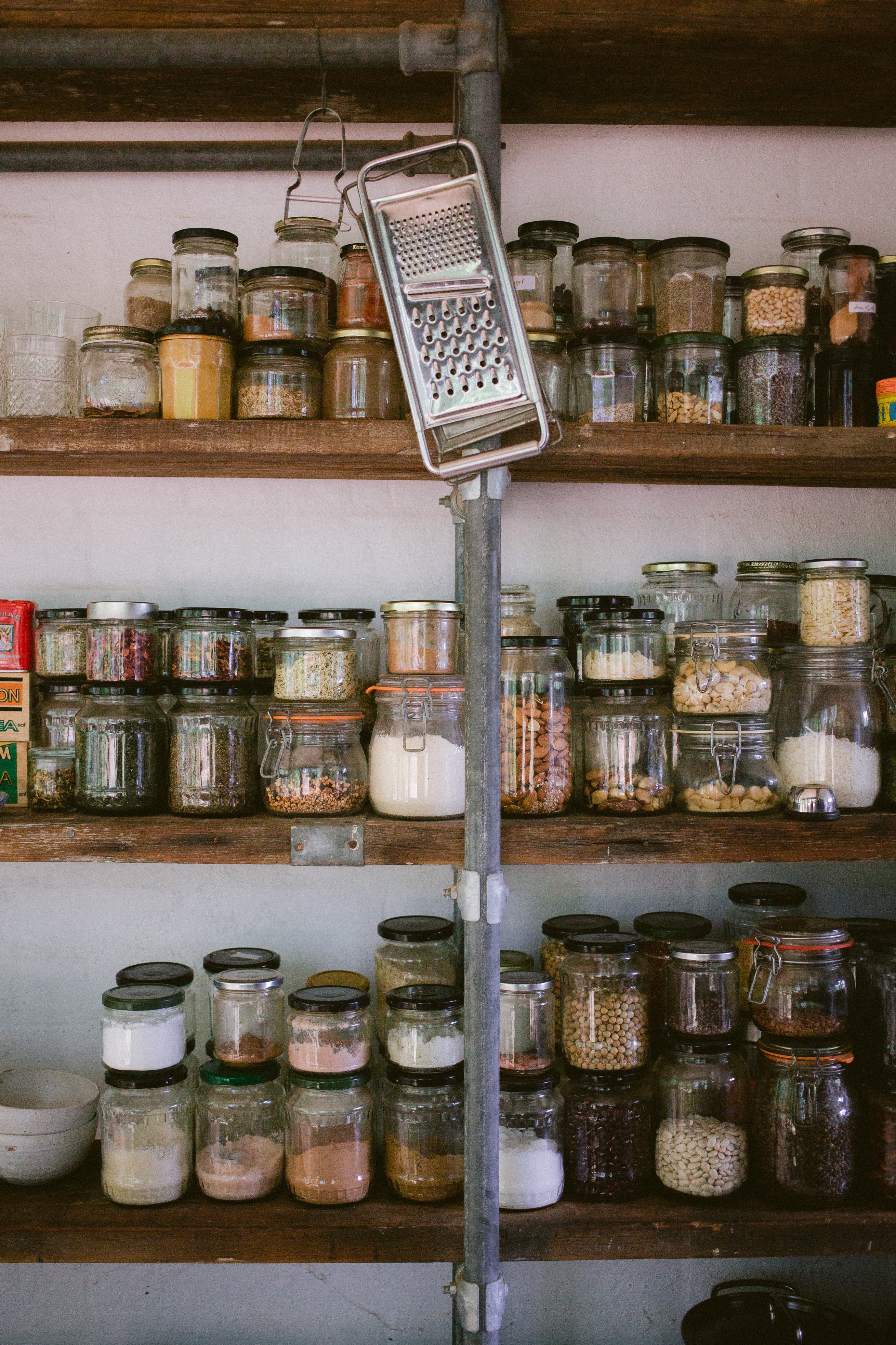 Photo of assorted jars with seeds and powders on rustic wooden shelves. In the center a metal pole from which hang a potato peeler and a grater. Your pantry will never start like this: you will build it slowly, picking up new ingredients as you learn. Same for other skills!